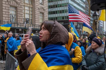 Foto de El alcalde Adams hace comentarios en la ceremonia de izamiento de la bandera para Ucrania. 24 de febrero de 2024, Nueva York, Nueva York, Estados Unidos: Una mujer envuelta en banderas ucranianas registra el izamiento de la bandera en una ceremonia de izamiento de la bandera para Ucrania - Imagen libre de derechos