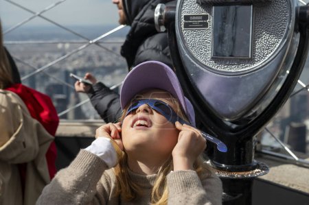 Téléchargez les photos : Solar Eclipse 2024 à New York. 08 avril 2024, New York, New York, USA : Nika, neuf ans, porte une paire de lunettes de protection pour voir l'éclipse solaire partielle depuis le pont d'observation du 86ème étage de l'Empire State Building - en image libre de droit