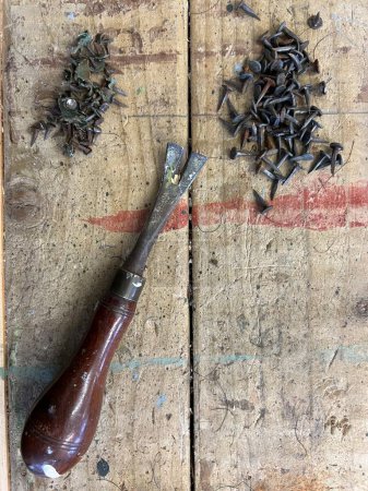 Close up of upholstery tool for removing tacks and nails with the vintage wood handle tool with metal hook on wooden work bench with piles of nails removed from chair upholstery renovation project