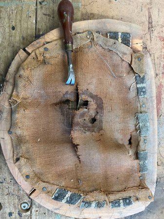 Téléchargez les photos : Close up of upholstery on vintage antique chair wood base of broken frame cotton webbing held tacks torn hessian material and tack nail removal metal vintage tool being re-made on work bench surface - en image libre de droit