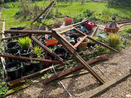 Garden landscape showing greenhouse being taken down with broken frame of wood posts on the ground with greenery, grass lawn, gravel pathway and plant pots and red watering can in early Summer sun day