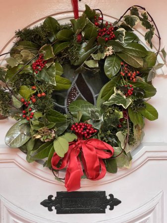 Téléchargez les photos : Gros plan de décoration de couronne de Noël avec beau houx vert, lierre, avec des baies, feuilles de laurier gui et arc rouge sur fond de porte peint en blanc - en image libre de droit