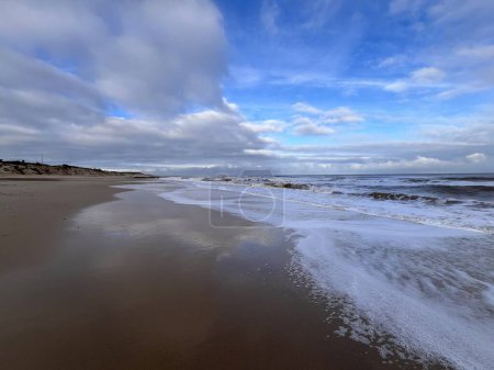 Téléchargez les photos : Belle vue sur le paysage de la plage de sable sauvage magnifique avec des vagues blanches reflètent les nuages dans l'eau de l'océan contre le large rivage de la mer et gris ciel bleu dramatique avec des nuages gris blanc pas de gens Journée d'hiver - en image libre de droit