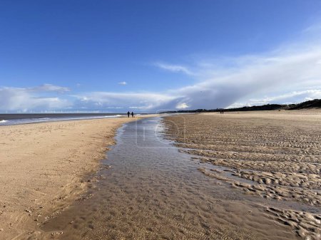 Foto de Impresionante hermoso paisaje de costa de playa de arena blanca en la marea baja río de entrada en la orilla al horizonte con pareja caminando cielo azul mar tranquilo y nube blanca Primavera frío día fresco en Winterton Norfolk uk - Imagen libre de derechos