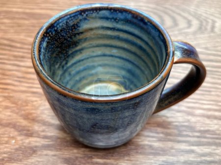 Close up of hand made ceramic mug the blue glazed empty rustic crafted cup with handle sat on wood kitchen surface interior ready to serve coffee