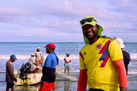 Foto de Cartagena, Bolívar Colombia - 1 de febrero de 2024: En primer plano, un pescador negro sonríe a la cámara y al fondo, un barco con motor fueraborda y sus compañeros arreglando sus herramientas, después de la pesca diaria - Imagen libre de derechos