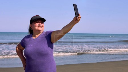Portrait d'une femme âgée et épaisse prenant un selfie sur la plage avec l'immensité de la mer derrière elle