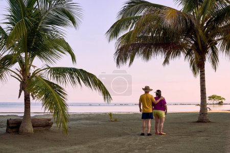 Ein Paar steht am Strand mit Blick aufs Meer, beide in Strandkleidung; er trägt einen "Vueltiao" -Hut und sie ein Visier. Sie stehen mit dem Rücken zu uns, zwischen zwei Palmen