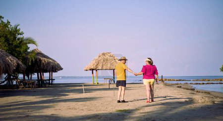 An elderly couple holding hands, dressed in beach attire; he wears a turned hat and she wears a large visor. They walk along the beach surrounded by thatched roofs and coastal trees, facing the sea