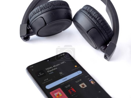 Foto de Lod, Israel - July 16,2023: Smartphone with TIDAL Music app play store page and wireless headphones on white background. Top view flat lay with copy space. - Imagen libre de derechos