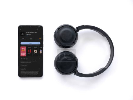Foto de Lod, Israel - July 16,2023: Smartphone with TIDAL Music app play store page and wireless headphones on white background. Top view flat lay with copy space. - Imagen libre de derechos
