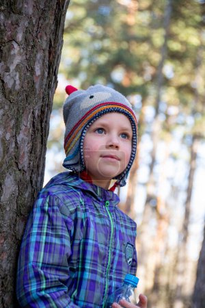 Boy spends happy time outdoors in forest enjoying autumn nature. A handsome little blond boy in a funny hat is looking into the distance near a big sprawling tree.