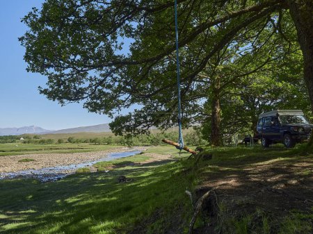 Photo for Offroad vehicle parking in the shade under a lush and green tree at the a river bank with iconic Ben Nevis mountain in the background. - Royalty Free Image