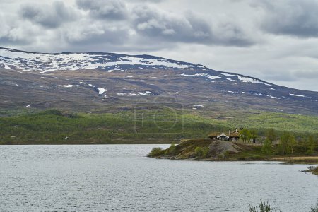 Téléchargez les photos : Maison typique en bois, chalet, le long des rives d'un lac dans les hauts plateaux Jotunheimen en Norvège avec des montagnes enneigées en arrière-plan. - en image libre de droit