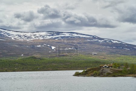 Téléchargez les photos : Maison typique en bois, chalet, le long des rives d'un lac dans les hauts plateaux Jotunheimen en Norvège avec des montagnes enneigées en arrière-plan. - en image libre de droit