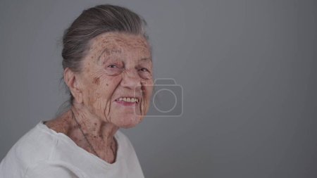 Seniorin 90 Jahre alt lächelt, lacht und zeigt eine Prothese auf grauem Hintergrund. Ältere Frau mit tiefen Falten und grauen Haaren glücklich in einem gut gelaunten Studioporträt, das in die Kamera blickt.