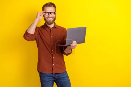 Photo of satisfied positive glad man blond beard dressed burgundy shirt hold laptop touch glasses isolated on yellow color background.