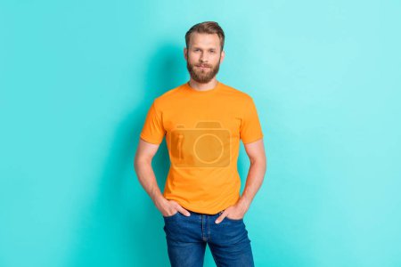 Portrait of satisfied confident handsome guy with blond beard wear orange t-shirt hold arms in pockets isolated on teal color background.