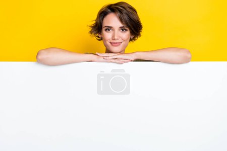 Photo of pretty positive lady hides behind empty space card blank recommend visit shop stylish clothes isolated on yellow color background.