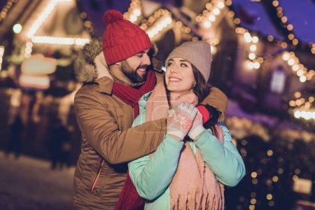 Foto von romantischen jungen Liebhabern Umarmung Look tragen Mütze Jacke Schal Handschuhe auf Neujahr Messe draußen.