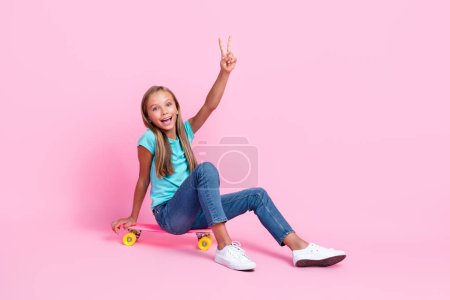 Foto de cuerpo entero de niño bastante pequeño emocionado usar turquesa camiseta monopatín que muestra v-signo de espacio vacío aislado de color rosa fondo.