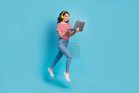 Foto de longitud completa de funky chica dulce vestido rojo camiseta auriculares saltar mecanografía dispositivo moderno aislado color azul fondo.