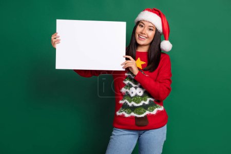 Photo of overjoyed pretty chinese woman wear red hat with ugly xmas pullover hold white paper banner empty space isolated on green color background. Poster 622415816