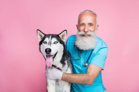 Foto de feliz alegre hombre mayor doc vestido uniforme azul abrazando perro husky sonriendo aislado color rosa fondo.