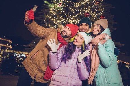 Photo of group excited positive buddies hold telephone make selfie portrait evening lights garland tree outside.