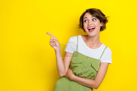 Photo of young pretty woman wear khaki overalls finger point empty space advert excited nice clothes market isolated on yellow color background.