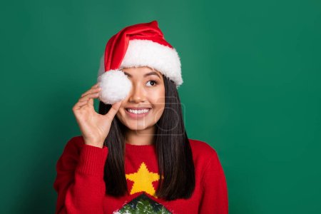 Photo of optimistic good mood girl with straight hairstyle dressed knit pullover cover eye santa hat isolated on green color background.
