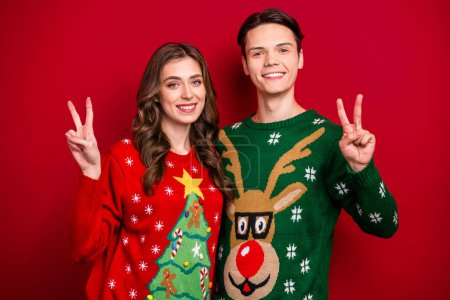 Photo of positive couple wife husband wear ugly sweaters demonstrate v-sign rejoice time together weekend isolated on red color background.