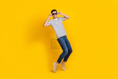 Full length photo of pretty funny guy dressed striped t-shirt dark eyewear having fun dancing isolated yellow color background.