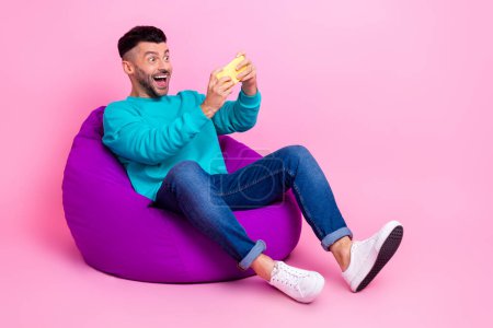 Foto de tamaño completo de joven guapo bolso de frijol de sentarse jugar juego móvil gamer desgaste ropa azul de moda aislado en el fondo de color rosa.