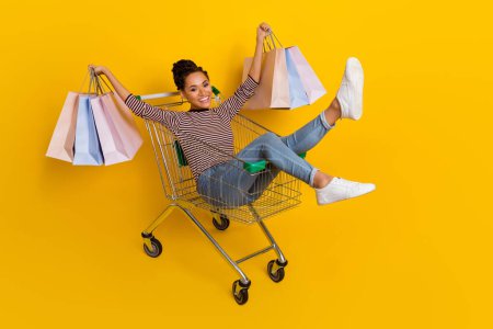 Full body portrait of excited overjoyed person hands hold packages sit trolley isolated on yellow color background.