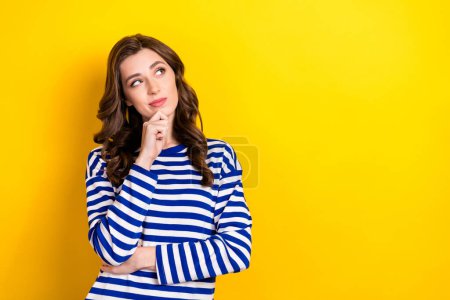 Photo portrait of pretty young girl look skeptical empty space consider dressed stylish striped outfit isolated on yellow color background.