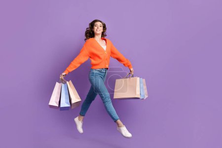 Full length photo of cute pretty woman dressed orange jumping high holding bargains empty space isolated purple color background.