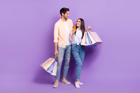 Ganzkörperporträt zweier positiver Menschen, die Boutique-Taschen in der Hand halten, sehen sich am Telefon isoliert auf lila Hintergrund an.