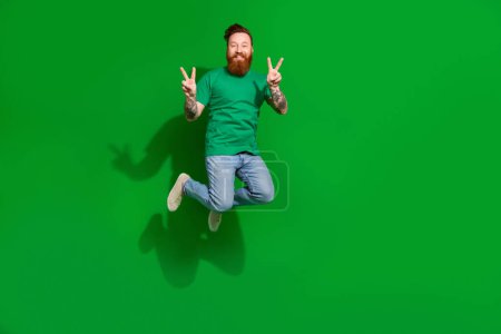 Full length portrait of overjoyed cheerful man jumping demonstrate v-sign empty space isolated on green color background.