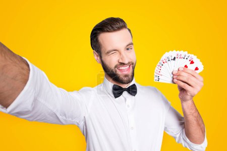 Self portrait of playful flirting croupier shooting selfie on front camera winking with eye demostrate present set of cards, isolated on grey background, having leisure video-call.