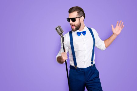 Porträt der hübschen attraktiven Sängerin in blauer Hose mit Hosenträgern und schwarzer Brille, singender Hit mit offenem Mund in Mikrofon-Geste mit isolierter Hand auf grauem Hintergrund.