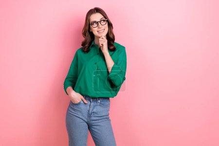 Portrait of good mood girl with curly hairstyle wear green shirt arm in pocket touch chin look empty space isolated on pink background.