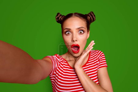 OOPS Self portrait of cute, trendy and shocked woman with bun hairdo wide open eyes mouth shooting selfie on front camera isolated on red background.