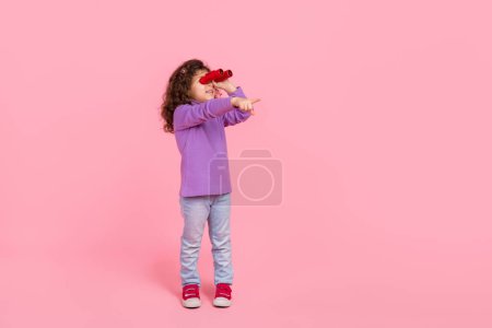Full length photo of excited little child dressed purple pullover watching binoculars pointing empty space isolated pink color background.