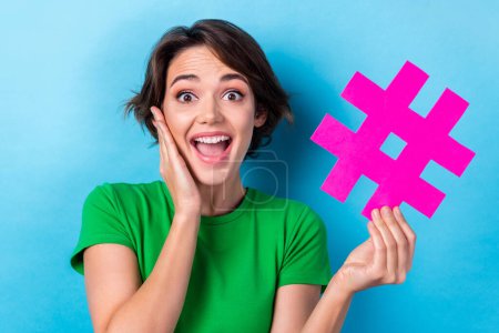 Portrait of surprised girl young age touch cheeks hold purple paper popularity hashtag symbol social media ad isolated on aquamarine color background.