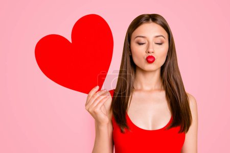 Portrait of charming woman with big paper carton red heart in hand waiting a kiss isolated on yellow vivid background.