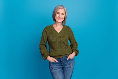 Photo portrait of attractive cheerful senior woman hold hands jeans pants pockets wear khaki sweater isolated blue color background.