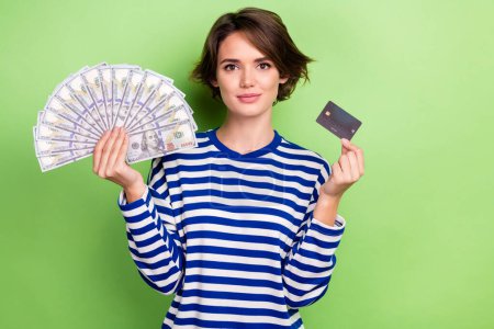 Foto von angenehmen hübschen Mädchen mit Kurzhaarschnitt gekleidet Matrose Langarm halten Plastikkarte Dollar isoliert auf grünem Hintergrund.