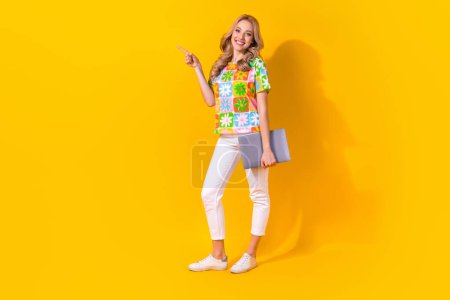 Full length photo of cheerful shiny lady wear flower print t-shirt holding device pointing empty space isolated yellow color background.