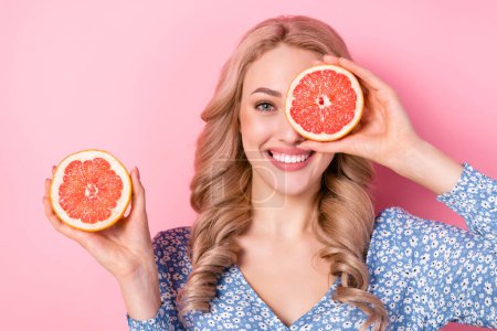 Portrait of young happy toothy beaming smile woman blonde beautiful hair cover eye half ripe grapefruit isolated on pink color background.